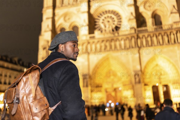 Young black man with a backpack and flat cap looking out while walking in front of notre dame cathedral at night, exploring the culture and architecture of paris