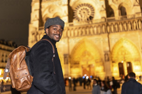 Young black man with a backpack smiling, visiting notre dame cathedral in paris france at night, representing travel, tourism, and european city exploration