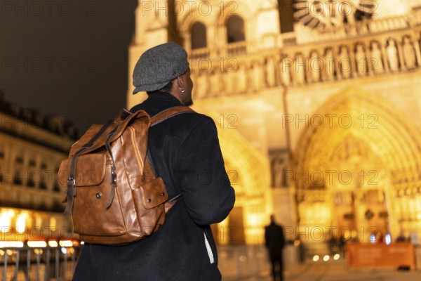 Solo man wearing a stylish cap and backpack, walking as a tourist during the night and looking at the illuminated architecture of a historic cathedral showcasing travel and exploration