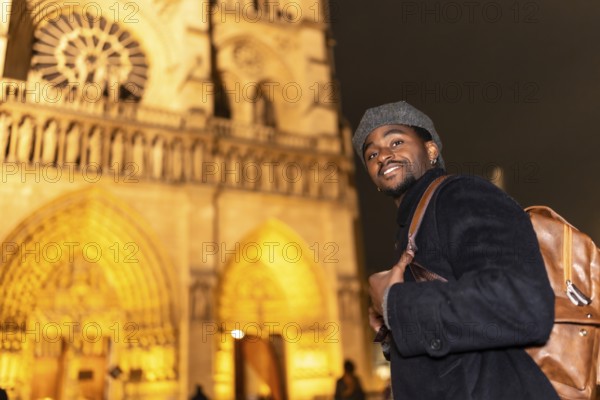 Young man smiling and wearing a backpack, exploring the historic architecture of notre dame cathedral at night, enjoying his holiday trip to the illuminated city