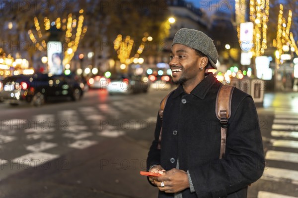 Smiling young black man wearing a hat and backpack, holding a smartphone while looking away, standing on a crosswalk in a busy city street illuminated by festive lights at night