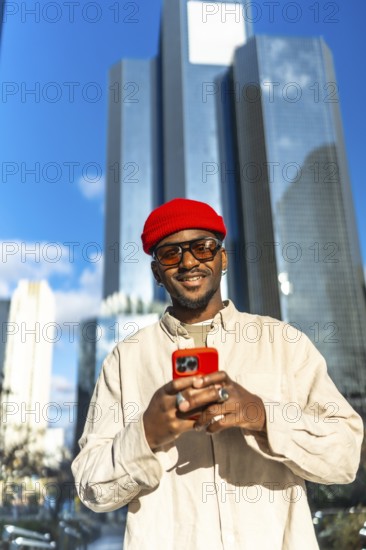 Stylish young black man wearing a red beanie and sunglasses, holding a bright red smartphone and smiling confidently in a modern city environment with skyscrapers under a clear blue sky