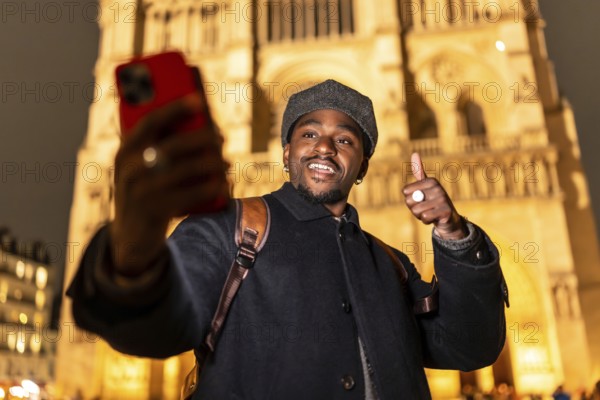 Young man smiling and giving a thumbs up gesture, taking a selfie with his smartphone in front of the illuminated notre dame cathedral in paris, enjoying city travel and nighttime tourism