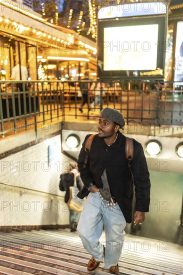 Young man wearing a beret, coat, and backpack ascending metro stairs in the vibrant urban environment of paris at night, with blurred lights from cafes and city signs in the background
