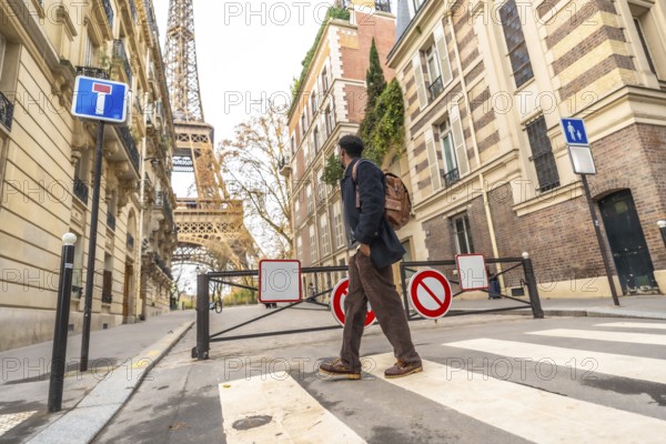Adult man traveling in paris, france, carrying a backpack and walking across a zebra crossing with the iconic eiffel tower and classic buildings in the background