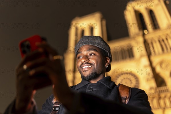 Young man enjoying a night street experience in paris, france, holding a smartphone and smiling while recording or taking a selfie in front of the illuminated notre dame cathedral