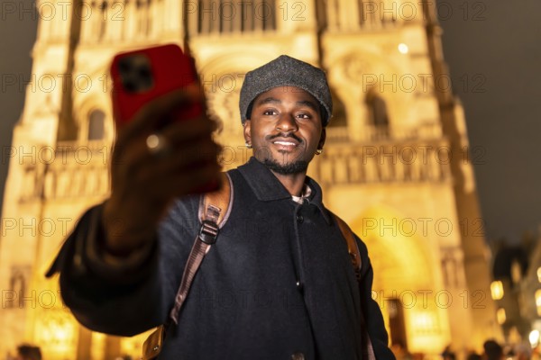 Young black man with a backpack smiling and taking a selfie with his smartphone in front of the illuminated notre dame cathedral in paris, enjoying a night out while traveling