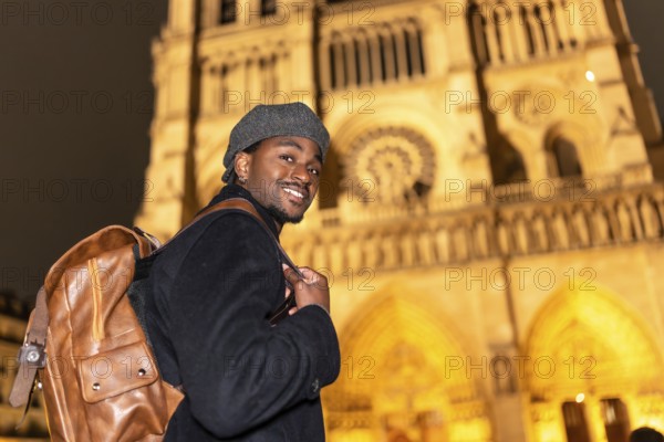 Young black man smiling, discovering french culture and history, standing in front of the illuminated notre dame cathedral in paris, celebrating travel and tourism