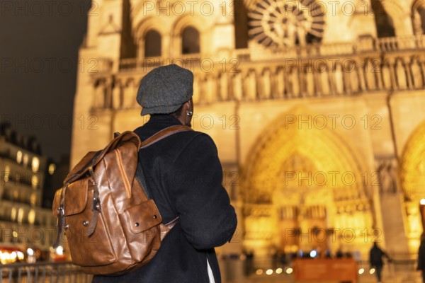 Tourist with a backpack and flat cap looking at the iconic notre dame cathedral facade, illuminated at night, capturing the concept of travel, discovery, and enjoying european landmarks