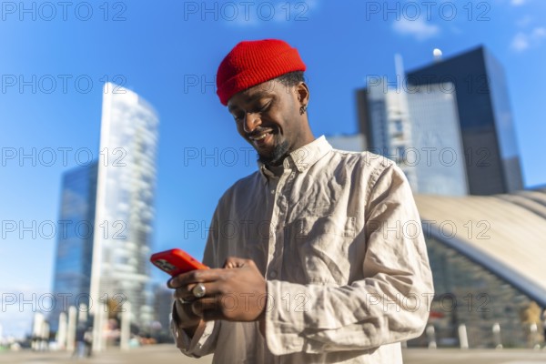 Young african american man smiling while typing on his smartphone outdoors in a sunny high tech urban environment with contemporary buildings in the background