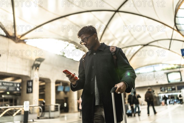 Indian businessman standing in a modern train station or airport terminal, looking down at his mobile phone while holding a rolling suitcase for travel