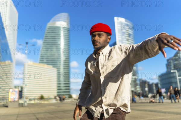 Black man wearing a red beanie and casual clothing moving rhythmically on a sunny day in an urban square with modern glass skyscrapers in the background, expressing joy and freedom
