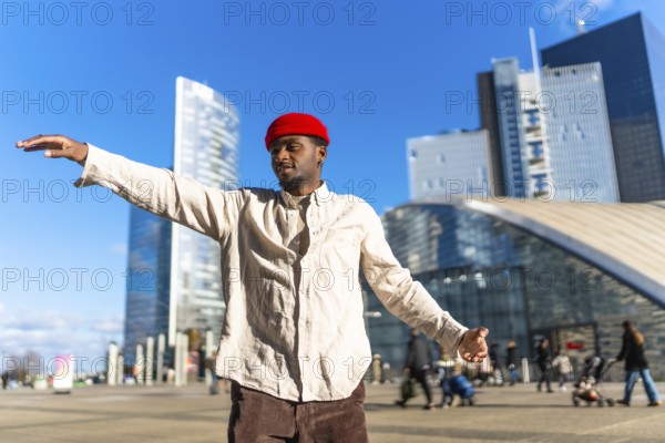Black man wearing a red beanie and light shirt expressing creativity through dancing, arms spread wide outdoors in a modern cityscape with skyscrapers