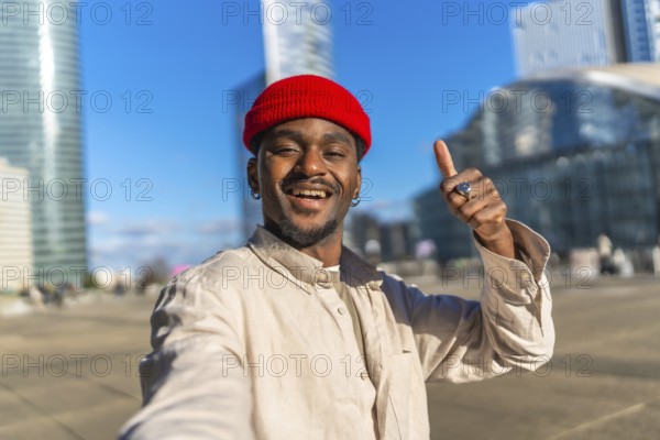 Young black man with a red beanie taking a close up selfie in an urban setting, smiling and giving a thumbs up gesture for success and approval on a sunny day
