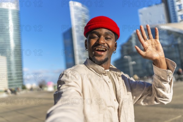 Young black man taking a selfie, smiling broadly and waving at the camera, showing a friendly gesture in a modern urban district with skyscrapers under a clear blue sky