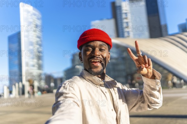 Young black man smiling and making a peace sign gesture while taking a selfie outdoors in a modern urban city with contemporary architecture under a bright blue sky