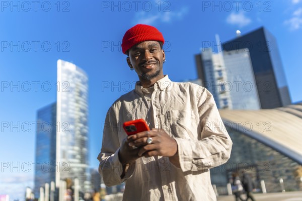 Young black man smiling at camera while using a red smartphone outdoors, standing in a contemporary urban environment with tall glass buildings under a vibrant blue sky during the day