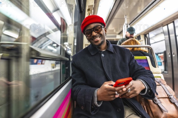Young man smiling directly at the camera while holding and using a red smartphone, sitting on a subway train during his daily commute and journey through the city