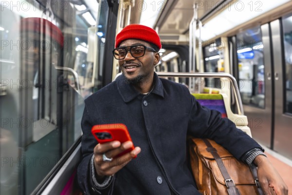 Young smiling african american man in a stylish coat and red beanie sits on a city train, holding a smartphone and browsing apps during a winter commute