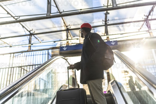 African american man traveling with a suitcase and backpack, riding an escalator upward in a modern train station or airport terminal with bright sunlight