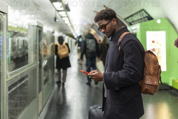 Young black man with a backpack and sunglasses standing on a subway platform, checking his smartphone while waiting for public transport in an urban environment