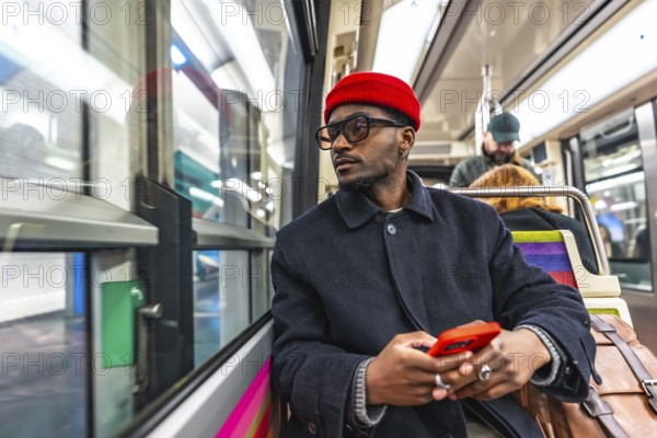 Stylish young black man wearing a red beanie and glasses sitting on a public transport train, holding a mobile phone and looking out the window during his daily commute