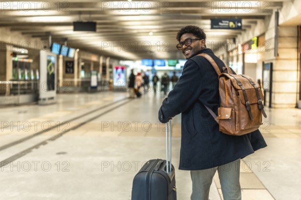 Smiling african man carrying backpack and pulling luggage, looking back while walking through a bright train station, representing business travel and commuting