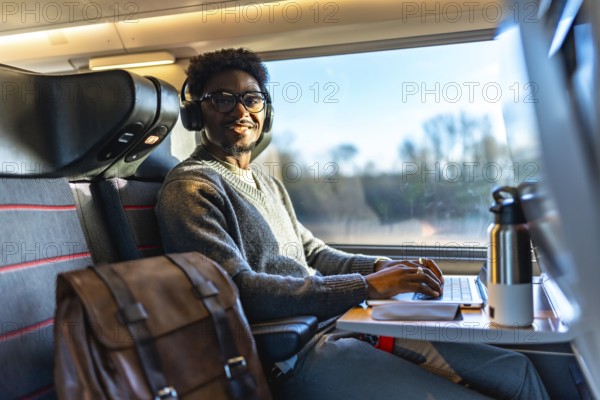 Young black man smiling while traveling on a train, wearing headphones and typing on his laptop, working remotely during a business trip or daily commute, focusing on productivity and connectivity