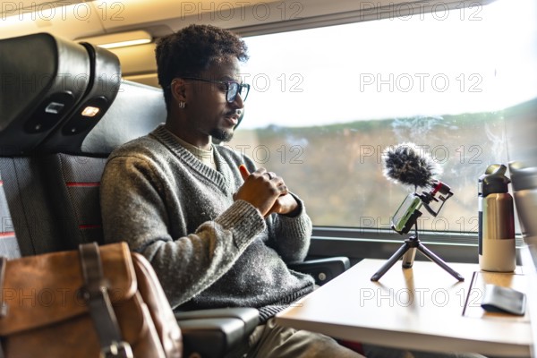 Young black man vlogging on a train, using smartphone and microphone on a tripod during a business trip remote work, focused content creator and digital nomad on the move