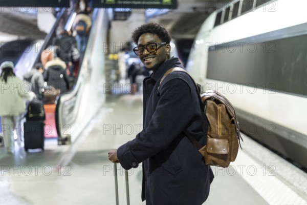 Smiling black man stands on a modern train platform with suitcase and backpack, ready for a business trip or commute confident, stylish, and prepared for urban travel and work