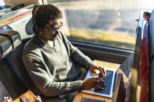Young black man focused on laptop on a train, wearing headphones and glasses while working remotely on a business trip, exemplifying digital nomad mobility and mobile office productivity