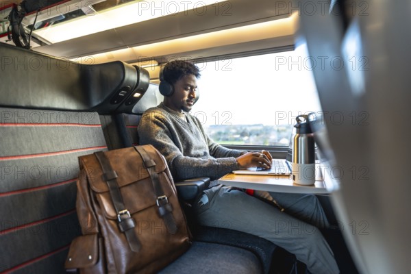 Young man wearing headphones working on a laptop while traveling by train, focusing on remote work and digital nomad lifestyle, enjoying a comfortable business trip