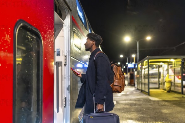 Young man standing on a train station platform at night, holding a smartphone and luggage, looking at the open door of a modern train, ready for a journey or commute