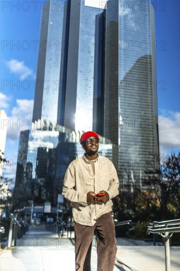 Young black man standing confidently in a modern city setting, wearing stylish casual attire and a red beanie, holding a smartphone with skyscrapers reflecting a bright blue sky