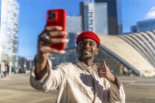 Young black man smiling while taking a selfie with a red smartphone and giving a thumbs up gesture, standing in front of modern architecture and skyscrapers under a blue sky