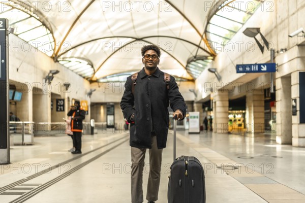Young businessman standing on a modern train platform, smiling confidently with suitcase and backpack, ready for a city business trip and about to board his train