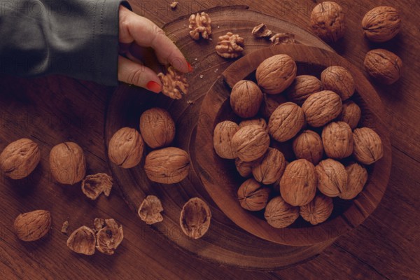 A woman's hand taking a walnut from a wooden bowl filled with walnuts. There are shells and walnuts scattered around