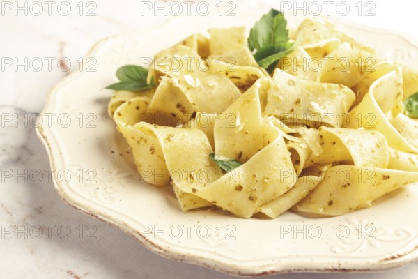 Fresh papardelle pasta, with basil and pesto sauce, is served, on a white plate, with a fork held by hand