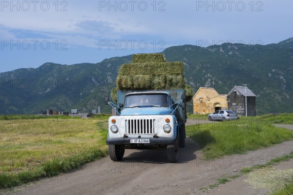 A blue truck with hay bales drives past a church, against a backdrop of mountains, vintage car, GAZ-53, near Odsun, Odzun, Saint Nshan Church, Lorikeet Province, Armenia