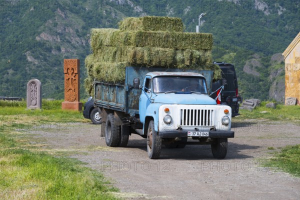 A blue truck with hay bales drives on a dirt road surrounded by mountains and fields, classic car, GAZ-53, near Odsun, Odzun, Lorikeet province, Armenia