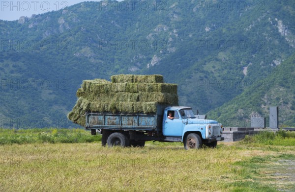 A blue truck with hay bales drives on a dirt road surrounded by green mountains, classic car, GAZ-53, Lorikeet province, Armenia