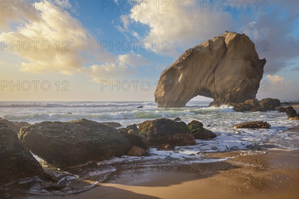 Penedo do Guincho, a large boulder rock arch at Praia da Santa Cruz, Portugal, with ocean waves and sandy beach on sunset