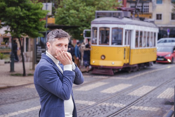Man musician in a blue blazer and white sweater playing blues on a harmonica with eyes closed, standing outdoors in Lisbon city street, Portugal with tram in background