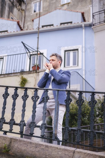 Man musician in a blue blazer and white sweater playing blues on a harmonica with eyes closed, standing outdoors in city street
