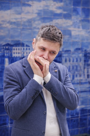 Man musician in a blue blazer and white sweater playing blues on a harmonica with eyes closed, standing outdoors in Lisbon city street, Portugal