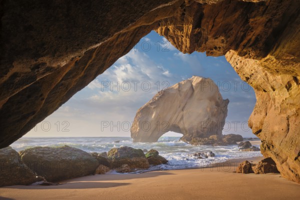 Penedo do Guincho, a large boulder rock arch at Praia da Santa Cruz, Portugal, with ocean waves and sandy beach as seen from grotto cave on sunset