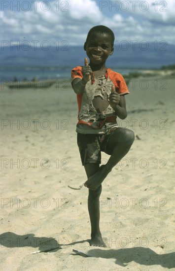 A cheerful local boy with a ripped shirt stands on the beach near Kande Beach on Lake Malawi and smiles at the camera, Malawi, Africa, July 2000, vintage, retro, old, historic