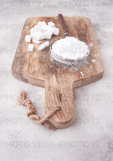 A strainer with powdered sugar, on a wooden cutting board, with pieces of white sugar