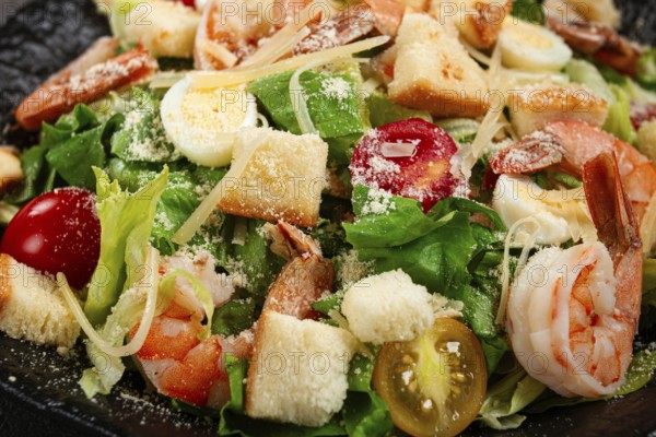 Caesar salad with tiger prawns, shrimp salad, lettuce leaves, quail eggs, cherry tomatoes, parmesan cheese, on a black plate
