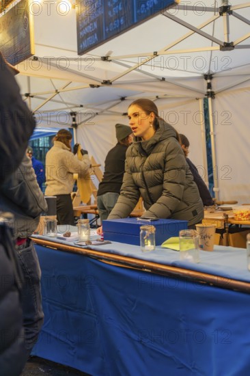 A woman in a winter coat sells goods at a market stall with a blue table, Aidlingen Christmas market, Böblingen district, Germany